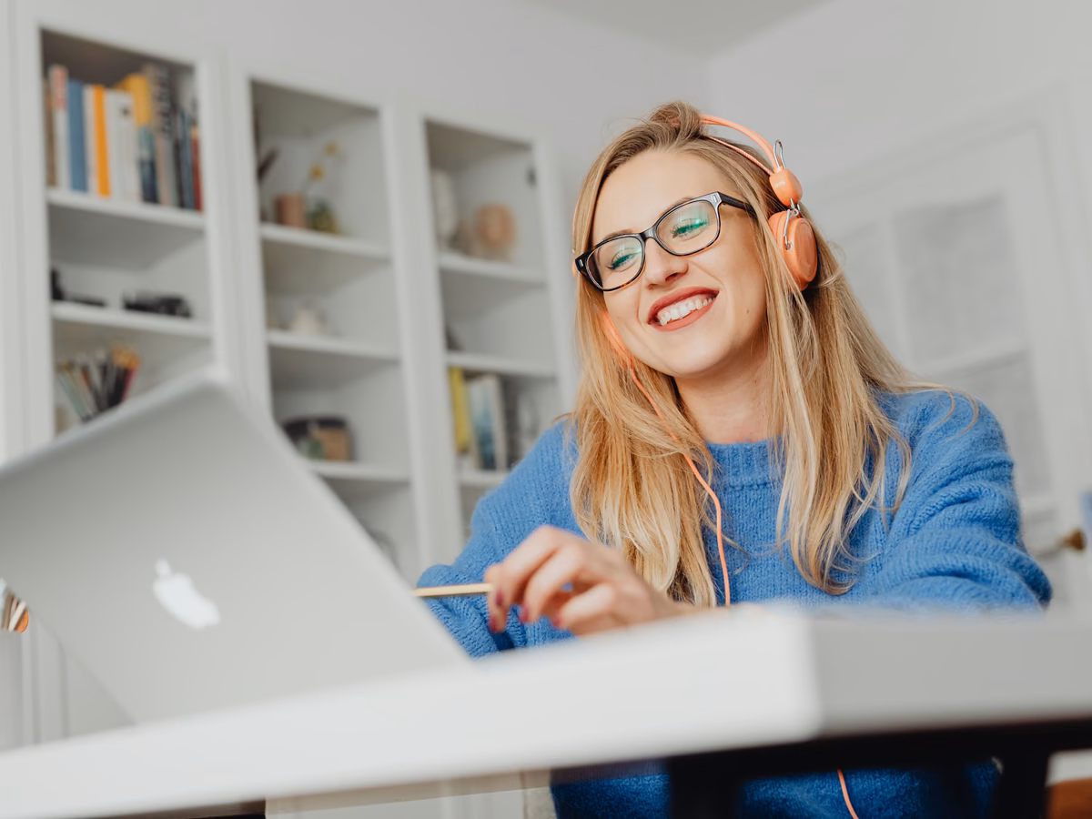 A smiling student in glasses and headphones following a live online class on her laptop