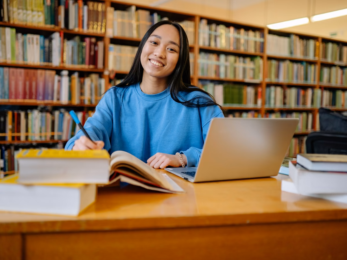 A smiling student working with her laptop and books in a library