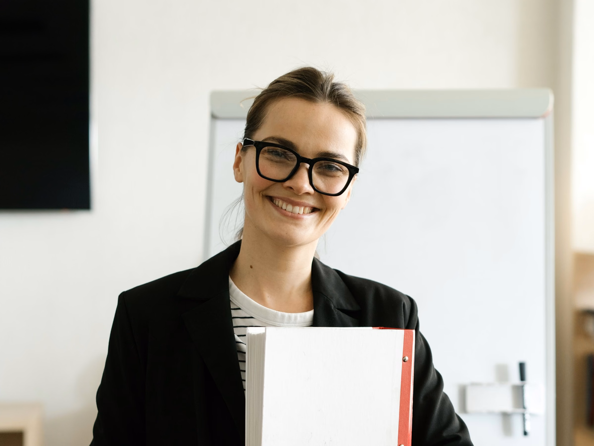 A smiling young teacher in glasses holding a folder in front of a classroom whiteboard
