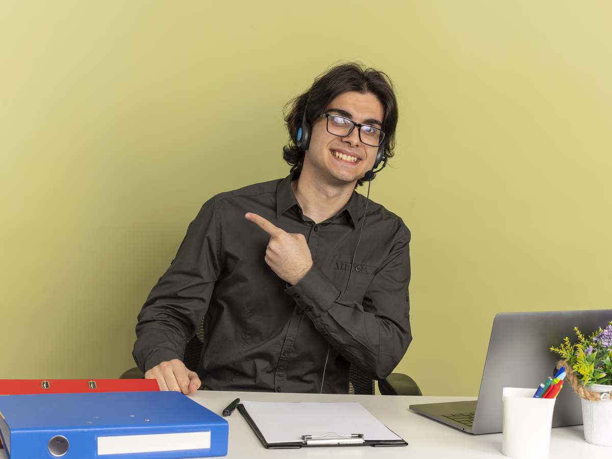 A smiling teacher in glasses and a headset at his desk with a laptop, a clipboard and folders, setting an online test